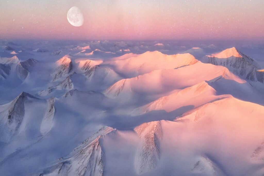 areal view of snowy mountain range with large moon in the background at what appears to be dawn