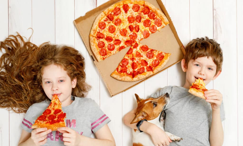 2 children lying on the floor with their dog next to a pepperoni pizza white background
