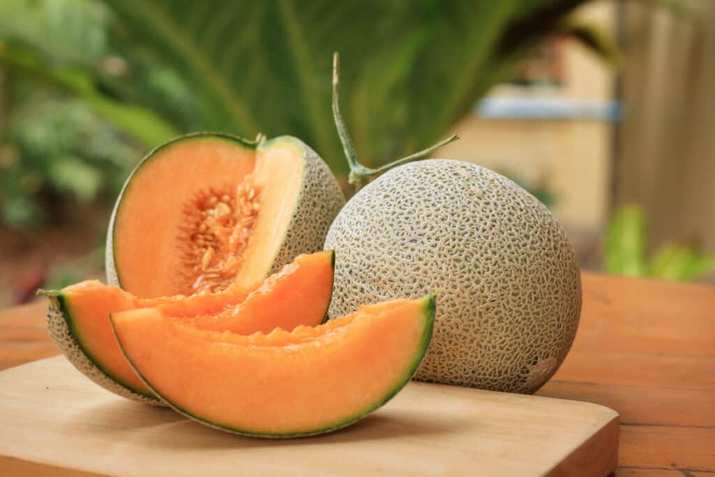 slices of orange cantaloupe on a cutting board with a whole cantaloupe in the background, along with some greenery.