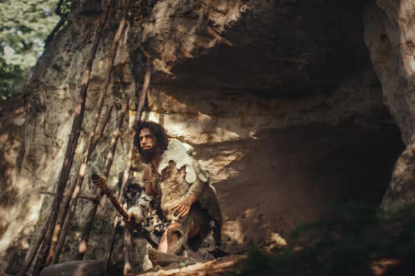 Primeval Caveman Wearing Animal Skin and Fur Holds Stone Tipped Spear Comes out of His Cave into Prehistoric Forest Ready to Hunt. Neanderthal Going Hunting into the Jungle