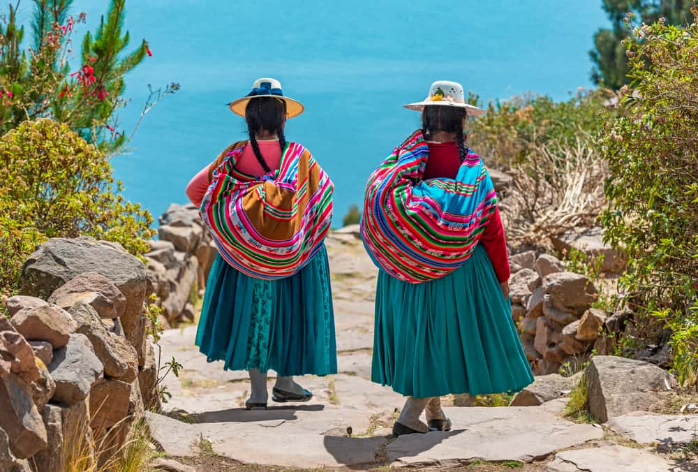 Arka planda Titicaca Gölü ile Isla Taquile (Taquile Adası) limanına giden yolda yürüyen geleneksel giysiler içinde iki yerli Quechua kadını, Peru.
