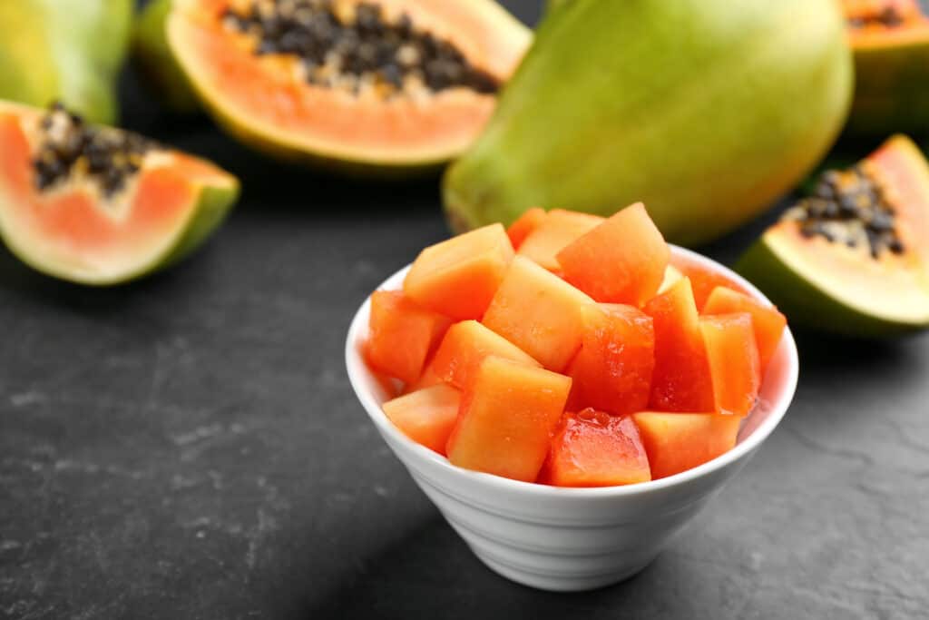 Papaya en rodajas en un tazón blanco con papaya entera y media detrás sobre una mesa negra