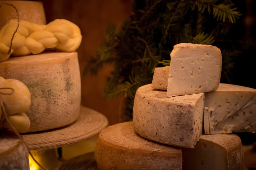 many different wheels and wedges of light colored cheese , stacked haphazardly on wooden cutting boards in a dark frame.