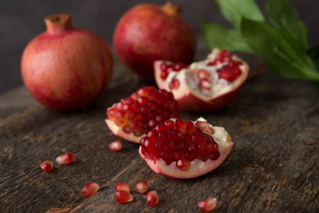 opened pomegranate with exposed fruits in from of two whole unopened pomegranates a wooden table