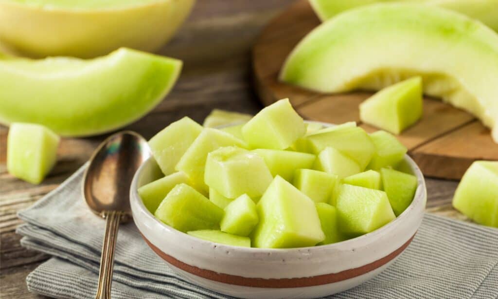 bite-sized pieces of honeydew in a ceramic bowl, resting on a napkin, along with a silver metal spoon atop a  wooden table. Honeydew wedges adorn the background.