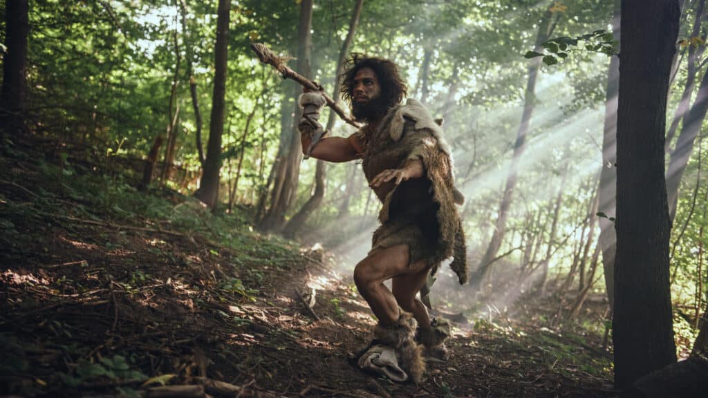 Primitive caveman in animal skin looking around with stone pointed spear exploring prehistoric forest for animal prey. Neanderthals went hunting in the jungle