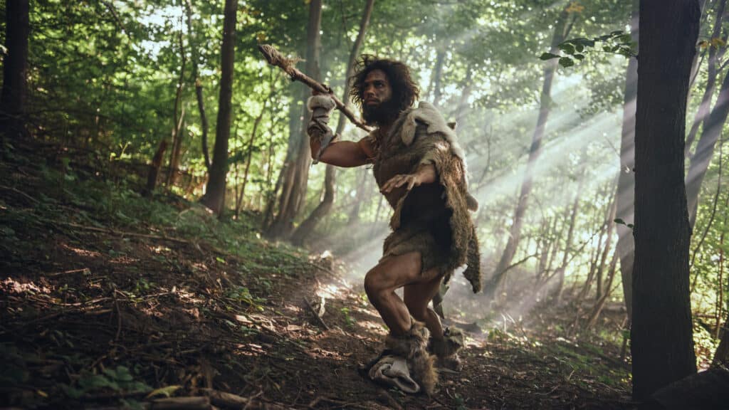 Primeval Caveman Wearing Animal Skin Holds Stone Tipped Spear Looks Around, Explores Prehistoric Forest in a Hunt for Animal Prey. Neanderthal Going Hunting in the Jungle