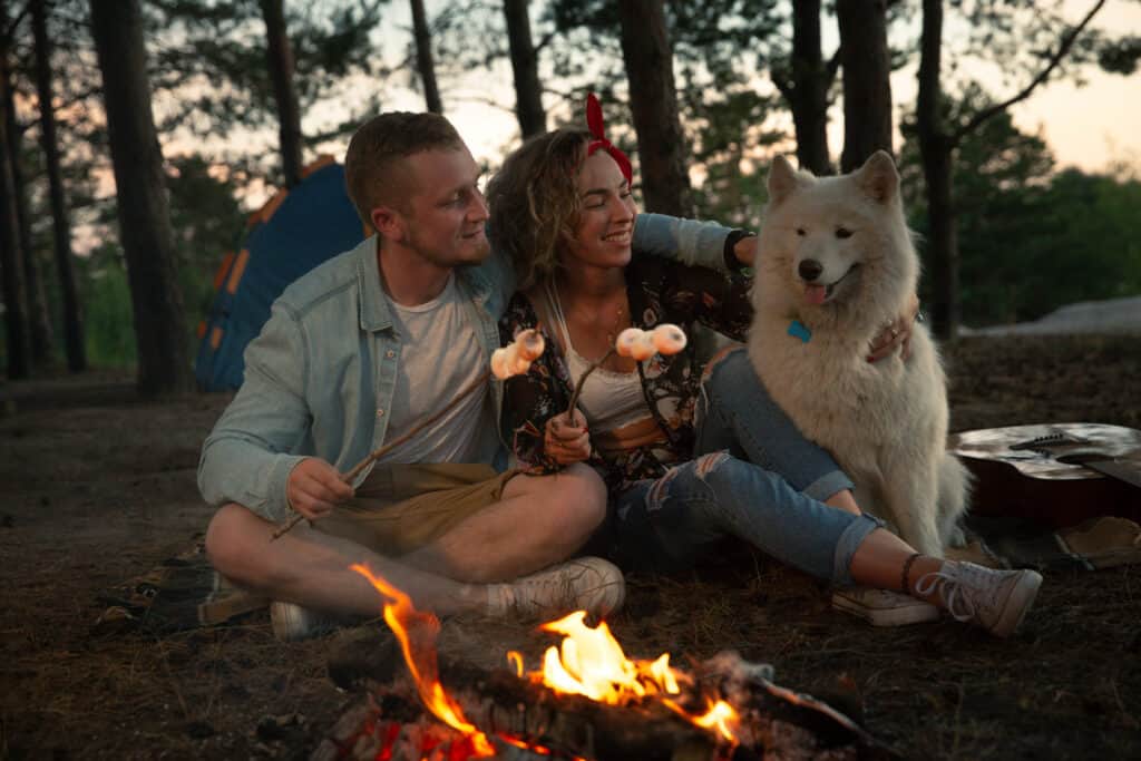 A guy with a girl and a dog by the fire to toasting marshmallows