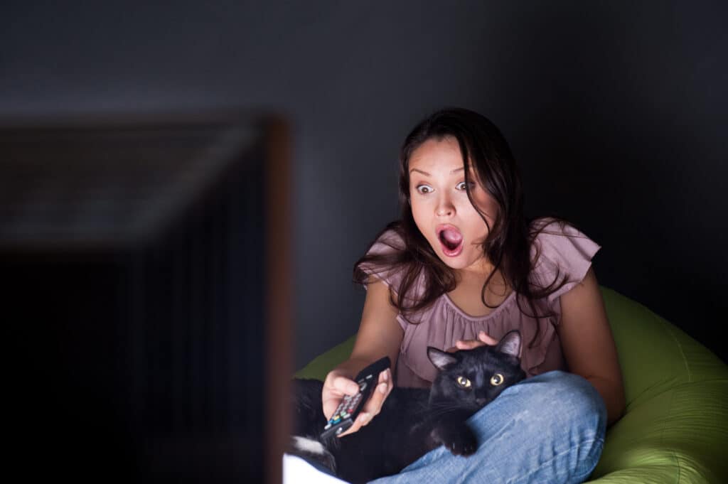 Young woman with black cat on her lap watching TV in the dark