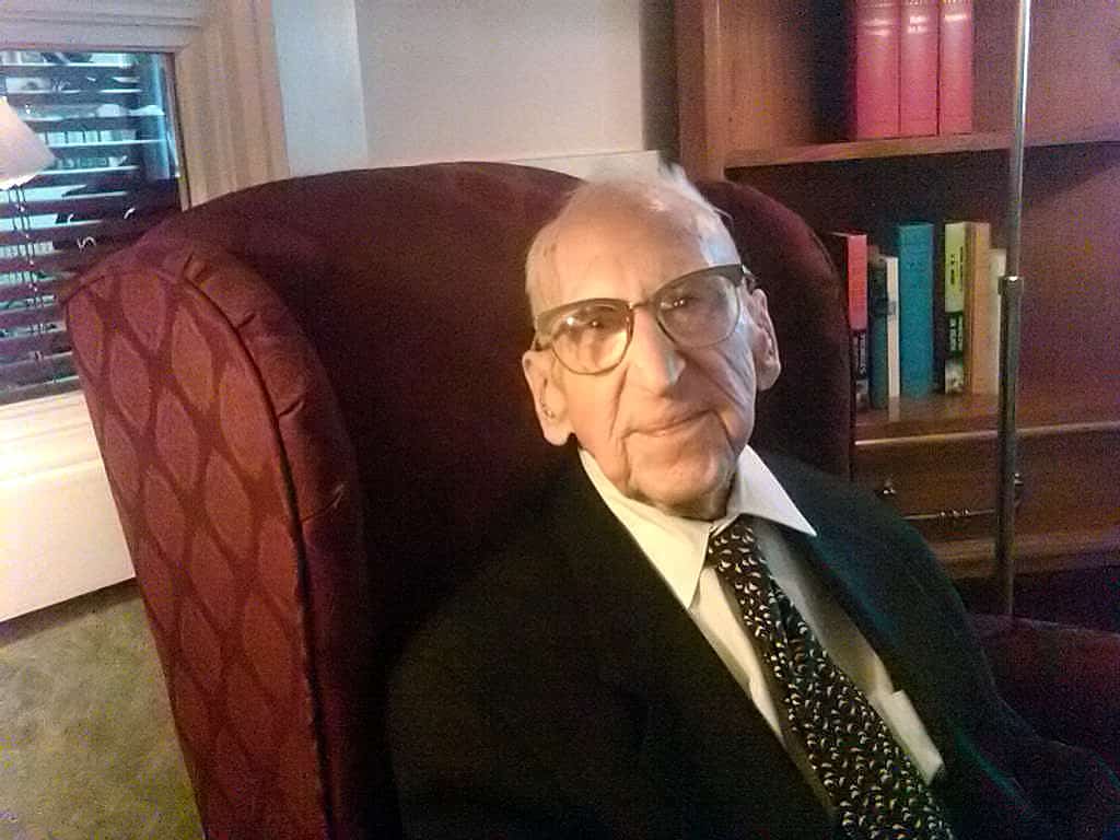 portrait of Walter Breuning once the world's oldest living man. He siseateid in a red upholstered chair . He is wearing a brome jacket with a light colored shirt and a brown tie. A bookshelf filled with colorful books is seen behind him. YJe chair's back is to a window/\.