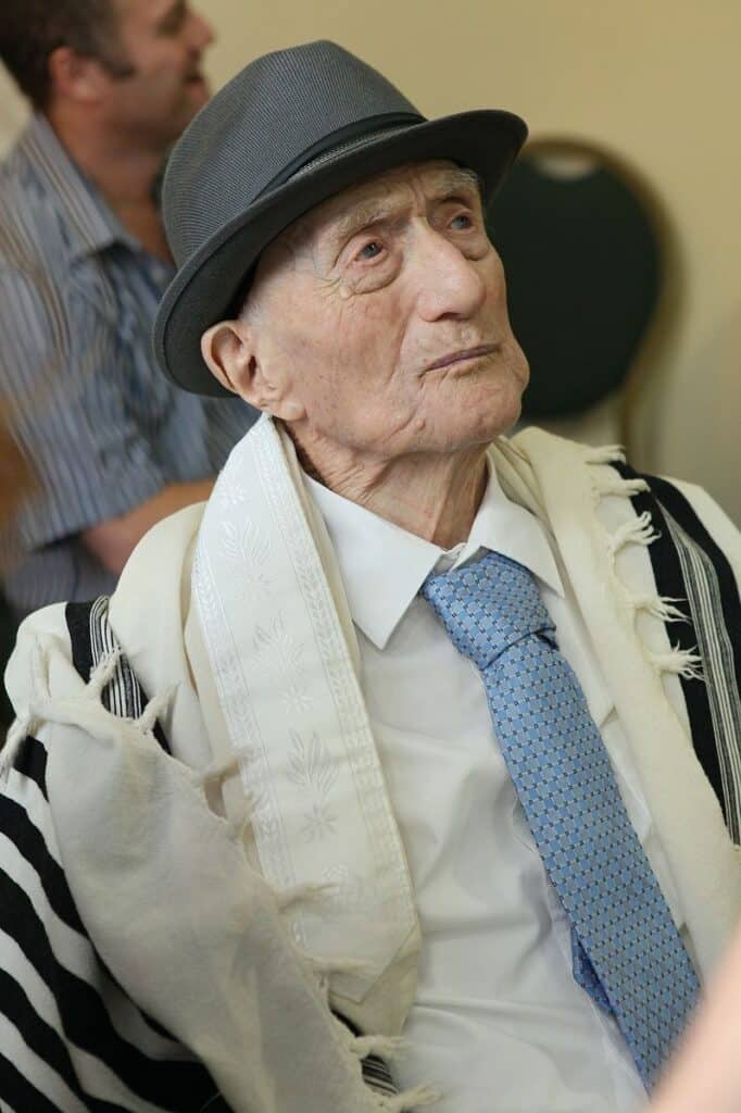 a portrait of Yisrael Kristal, once the world's oldest living man. Kristtal is light skinned, wearing a  dark grey fedora hat a prayer shawl, a white shirt and a light blue tie. 