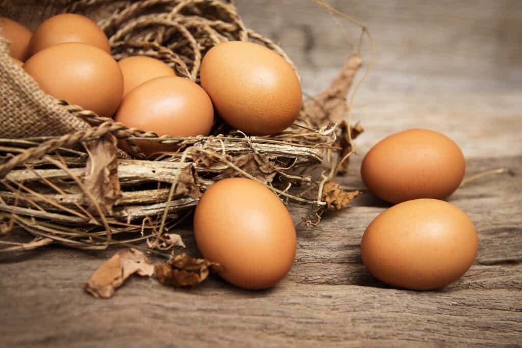 Three brown chicken eggs that appear to have rolled out of a basket onto and uneven wooden surface. The basket still contains at least six brown eggs. The basket resembles a bird's nest. it is Ade from twigs and leaves and has some associated burlap. 