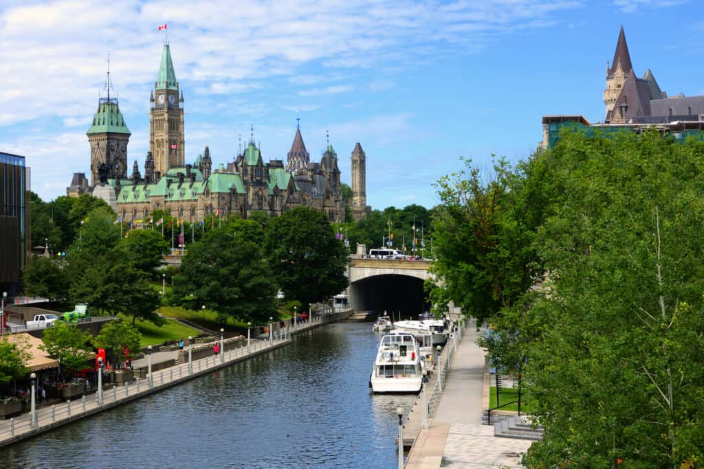 The Rideau canal in Ottawa, Canada in the summertime. Green trees flank the canal. two boats are anchored on the right side of the canal and Ottawa's parliament Hill can be seen in the background. An older building with turrets and mint toothpaste colored roof. 