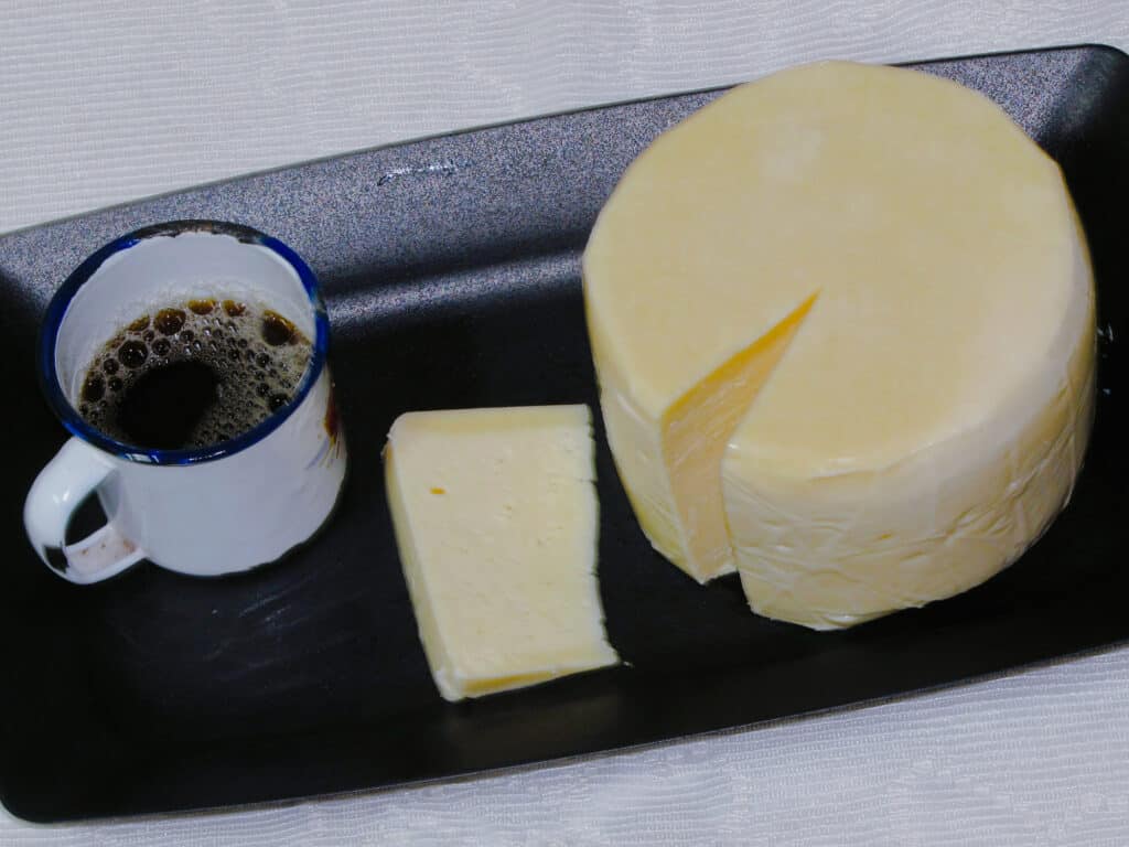Camera angle is high, looking down upon: a round wheel of very light yellow Serro cheese - it looks like butter. A slender wedge has been removed and lies next to the wheel/ A wheel used metal cup coated in white enamel and half filled with a dark brown liquid with tan bubbles on its surface, sits to the left of the wheel and the slice on a nondescript dark metal tray. against a white background