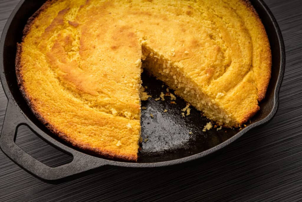 A cast iron skillet with cooked goldencornbread in it. One triangular wedge is missing, between 4-6 o'clock. The skillet is on a darn brown wood table. 