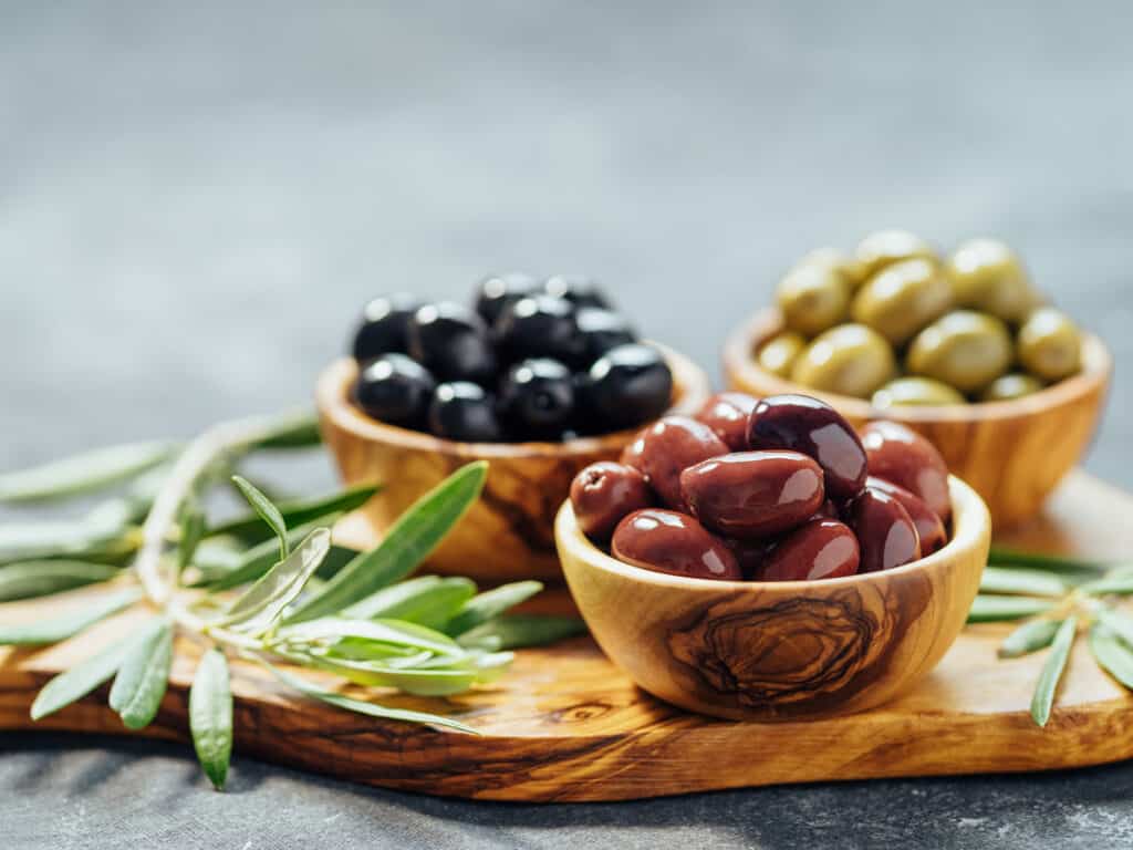 three Woden bowls, one each of ripe-black, kalamata, and green olives on a wooden cutting board adorned with rosemary.