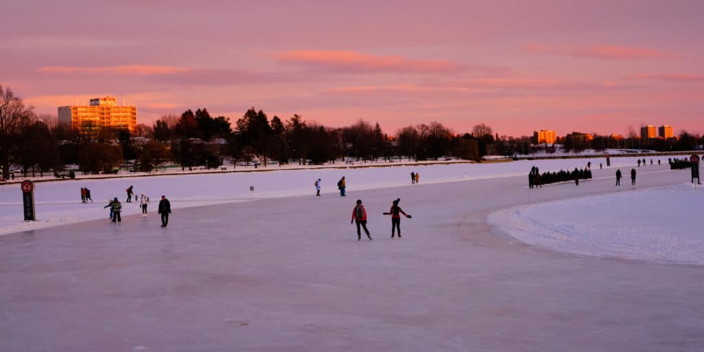 The Rideau Canal Skateway at twilight. no more than a dozen skaters are on the ice, against a pink sky with lavender accents. 