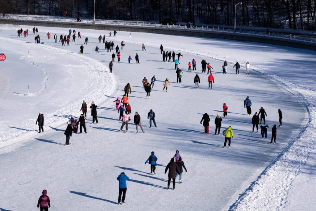 The world’s largest ice rink is the Rideau Canal Skateway in Ottawa, Canada. Close to 100 people in brightly colored and dark clothing, ice skating on a frozen, snow covered canal; they are skating counter-clockwise, moving toward the upper left corner of the frame. 