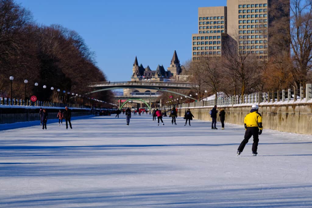The world’s largest ice rink is the Rideau Canal Skateway in Ottawa, Canada. 10 skaters can be seen in the frame, though there are more in the distance. The primary skater in the frame is wearing a bight yellow jacket and dark pants. Ottawa's Cityscape, modern and older building in background. One building five visible turrets. 