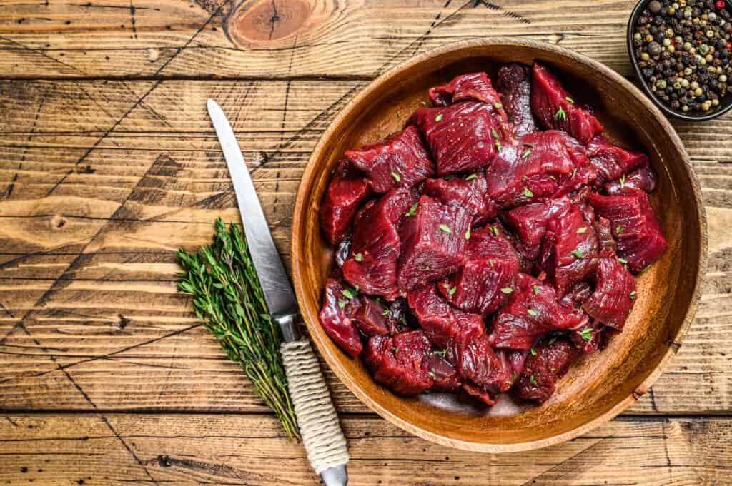 Raw cut wild venison meat for a goulash in a wooden plate. Wooden background. Top view. Copy space