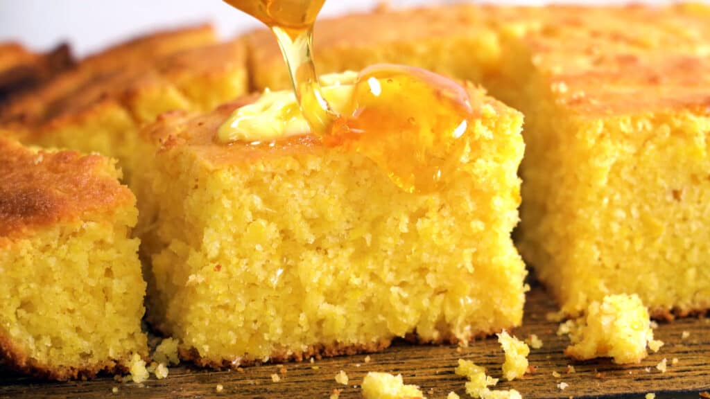 One full square piece of cornbread center frame flanked by two half pieces, one on either side, with more slices behind them. The central full piece is having honey poured on it .