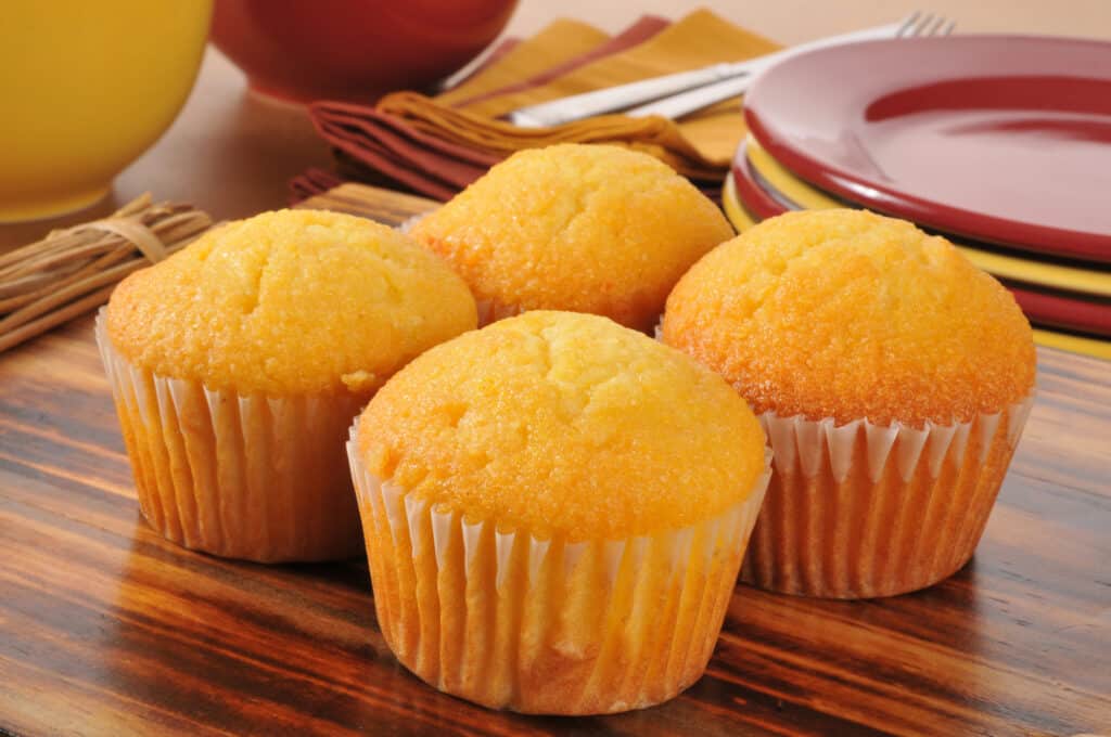 Four golden yellow cornbread muffins in white paper cupcake papers grouped  center frame on a woodgrain composite veneer table. a stack of four visible alternating red and yellow stoneware plates can be seen behind the plates, upper right. some yellow napkins with a silver metal fork and knife are seen to the left of the plates. Two large bowls, front yellow, back red, are upper left frame next to napkins.  