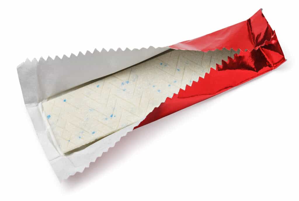 one stick of gum resting in an opened wrapper, which is shiny red foil on the outside and white on the inside. The wrapper has a pinked edge. The gum is off-white flecked with blue. white isolate background.