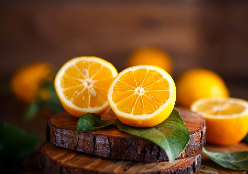 An orange Meyer cut in half, sitting atop green leaves, on a natural 1/2 inch slab of a pine tree trunk cutting board with another half of plus two whole  orange Meyer lemons. 
