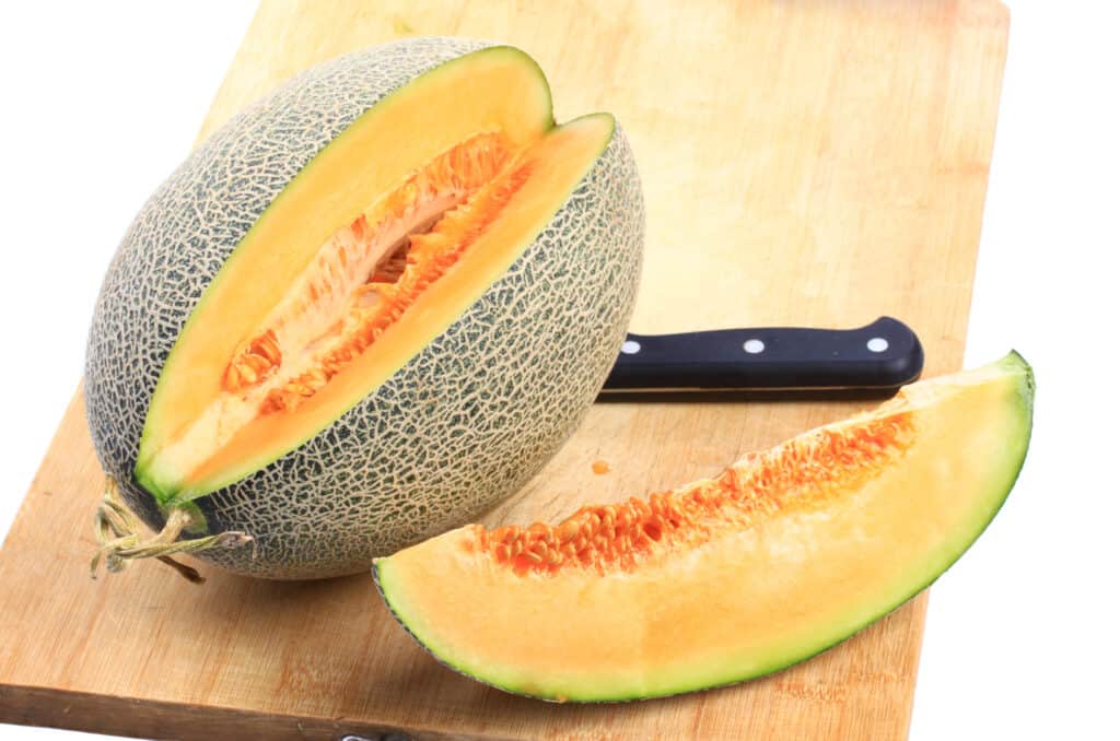 a hami melon ( looks like an oblong cantelpe, though the flesh is a bit lighter) on a cutting board with one wedge cut out, a knife resting on the far side of the cutting board. 