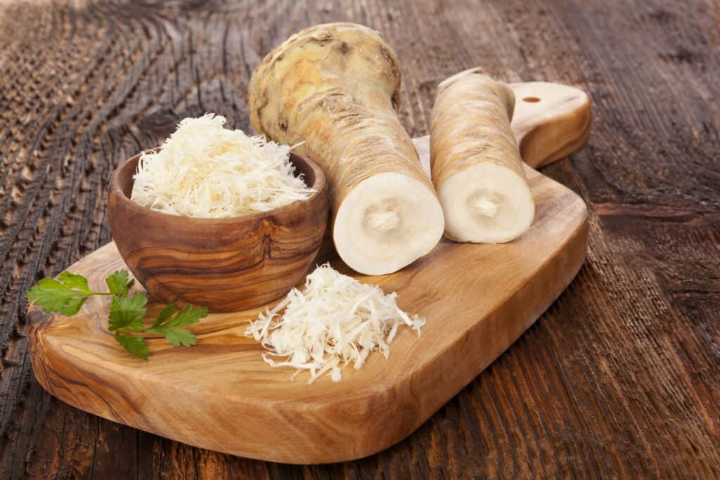 a bowl of grated horseradish sitting on a cutting board with two horseradish roots, a pile of freshly grated horseradish, and a sprig of parsley.