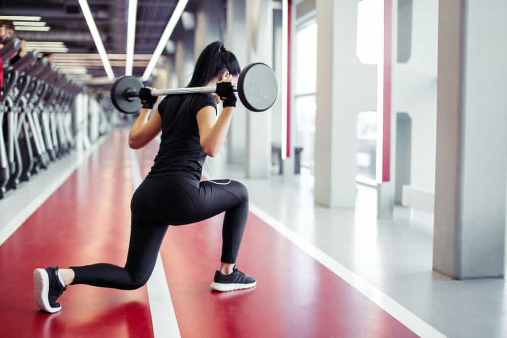 A female presenting human doing squat with a barbell with weights on each end across the back of her shoulders. She's is dressed in black leisyre attire, on knee resting a red track with a vertical white line running through the center.