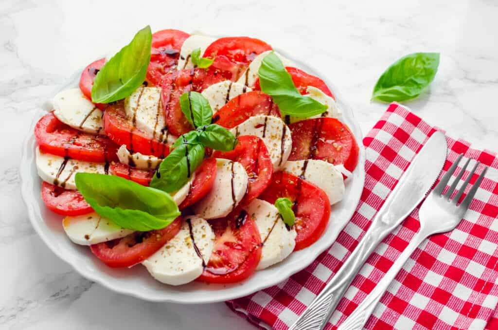 A caprese salad (Slices of red tomato and slices of fresh white mozzarella with a garnish of fresh sweet green basil leaves and drizzled with something brown -possibly balsmic in nature in a white ceramic bowl on a table with a white table cloth and a red gingham napkin upon which Sith a silver metal knife and fork. One basil leaf has been placed esthetically on the table.