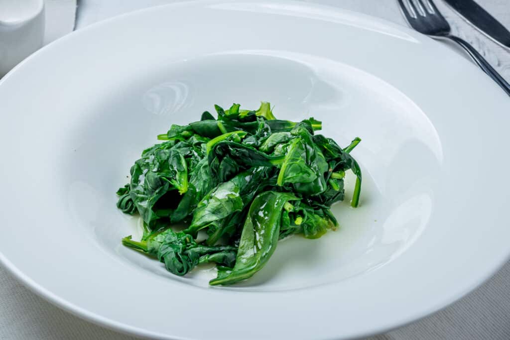 Center frame: A serving odf very green seemed spinach in a large white ceramic wide-rimmed bowl.A silver metal fork and knife are in the up right corner frame, to the right of the bowl. All on table with white table cloth.