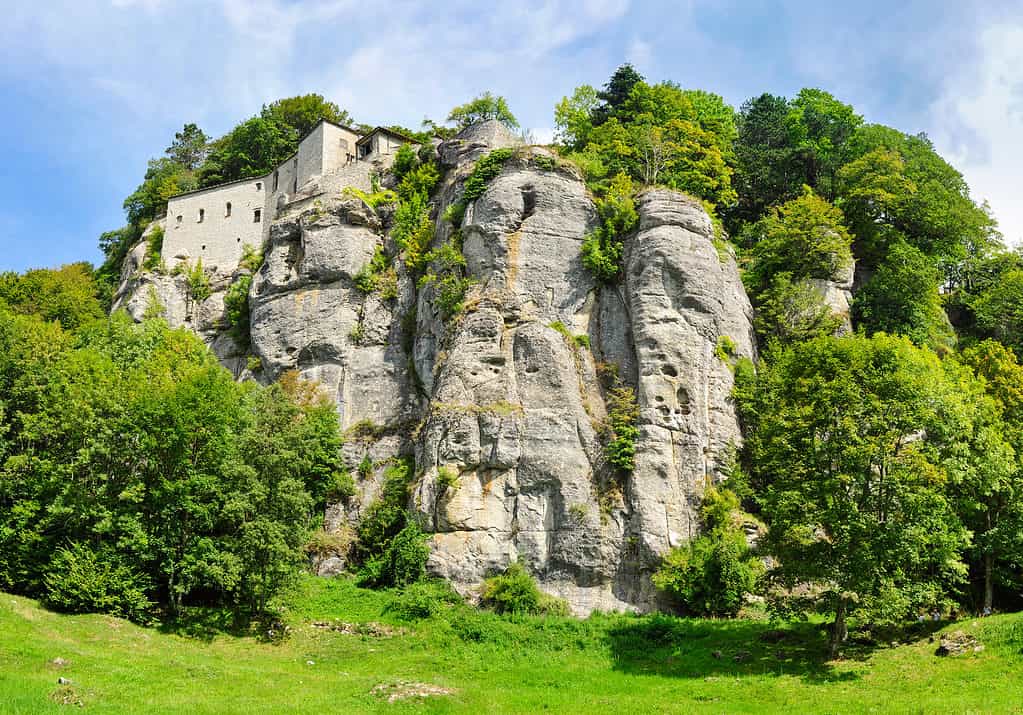 A white brick or stucco building ivisible upper left frame. It is perched onto of a large gray to brown rock outcropping tat is surrounded by greenery. There is grass in the foreground, frame left -right, and an opartly cloudy blue and white sky, upper frame- left to right.