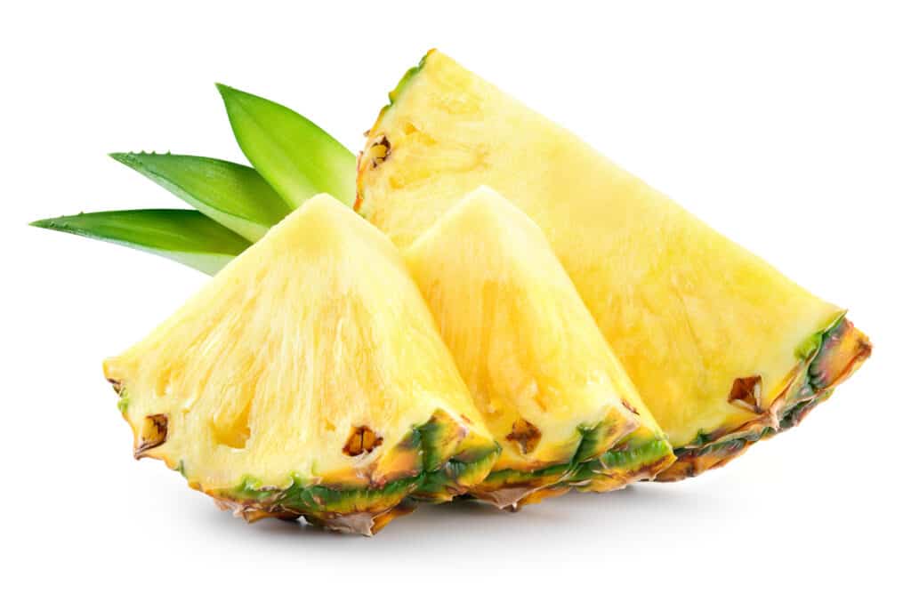 Center frame:two triangular wedges of fresh pineapple are seen in front of a semi-circle cut wedge. with three pineapple fronds against white isolate. The pineapple's flesh is light yello, ints rind/skin orange/brown. with dark green accents. The three fronds (leaves) are arranged together aesthetically. They are greener at the tip, becoming more yellow farther away from the tip.