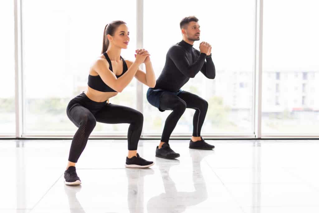 Female, front, and male,rear, dressed in black leisure wear, performing a squat, with their legs bent at 90 degree angle, their arms in front of their chests, clasped together. The floor is white and shiny, the background consists of three plate glass windows.
