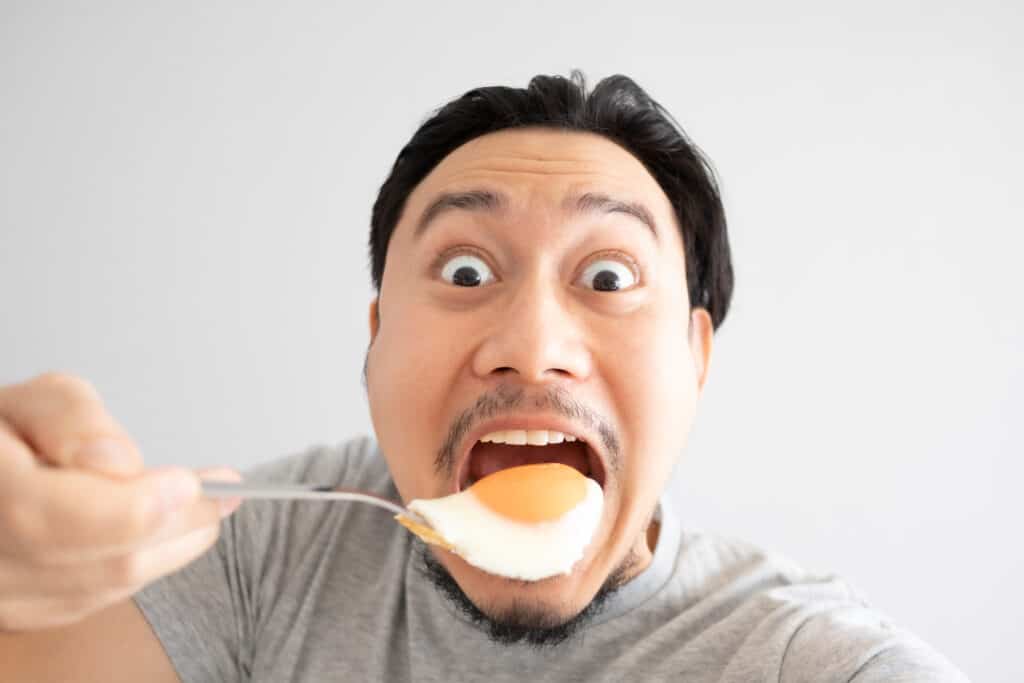 A light skinned man with a thin mustache wearing a grey t-shirt. His mouth is wide open as he prepares to fill it with an entire sunny side up chicken egg off of a silver metal utensil. The egg is white with a dark yellow center. 