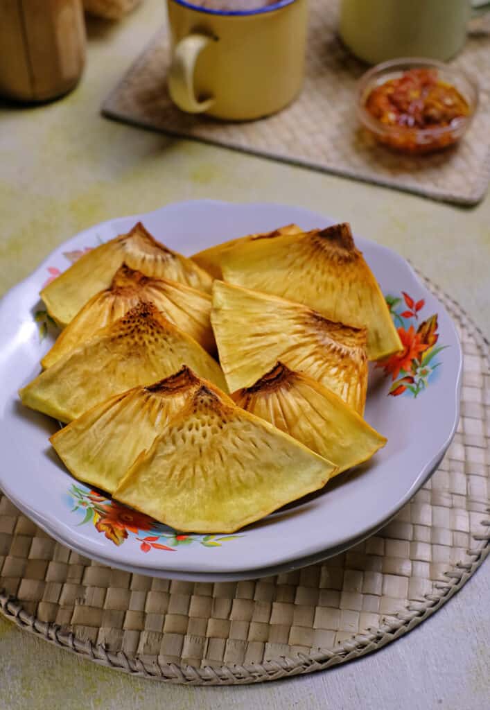nine thinly sliced triangles of fried, yellowish breadfruit on a porcelain plate with floral accents. atop a woven reed placemat. Background consists of a yellow ceramic mug with a cobalt rim and a small, clear glass condiment dish that contains a dark red sauce. Both the cup and the condiment dish are also on a woven placemat/.