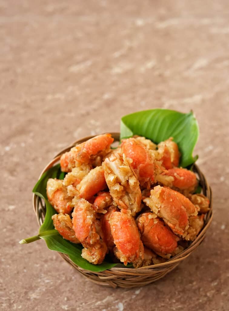 A woven, light brown, grass bowl filled with crispy sand fleas to be eaten. The small crustaceans are on top of a green leaf. The bowl sits on a taupe marble table.