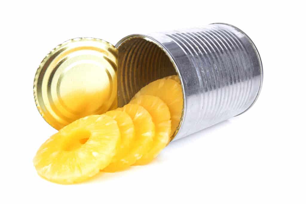 A silver tin cantered on its side, at a slight angle, with the bottom of the can, toward upper right corner of frame, and open top toward lower left frame. Four slices of pineapple are arranged as if the have slid out of the can, knocked over domino-style, with other slices still in the can seen behind them. The pineapple is golden yellow. The inside of the top of the can, which is still attached, is gold in color, and folded back , toward frame left.