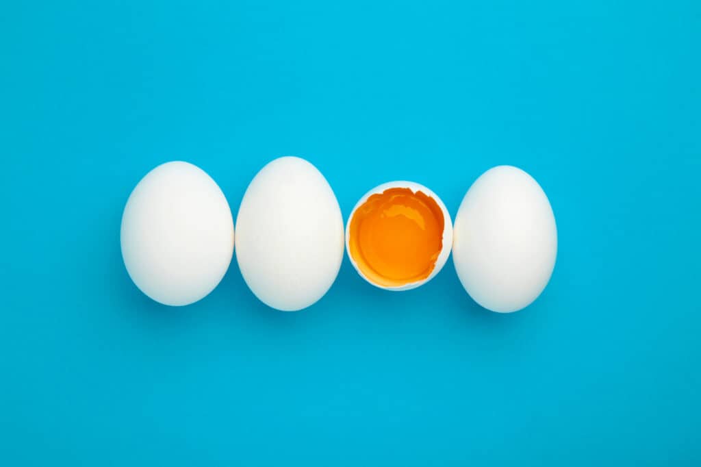 Center Frame: Four white chicken eggs against blue isolate. The third egg from the left is open, exposing an orangey-yellow yolk.