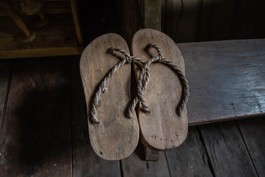 Center frame: Avery simple pair of rope and wooden sandals against a wooden background. The sandals resemble ancient flip-flops, with a loop of string attached to the wooden sole at the top for the toes to grasp, with one upside down V shape of rope attached at the heel end of the wooden sole on theft and the right. All natural colors. 