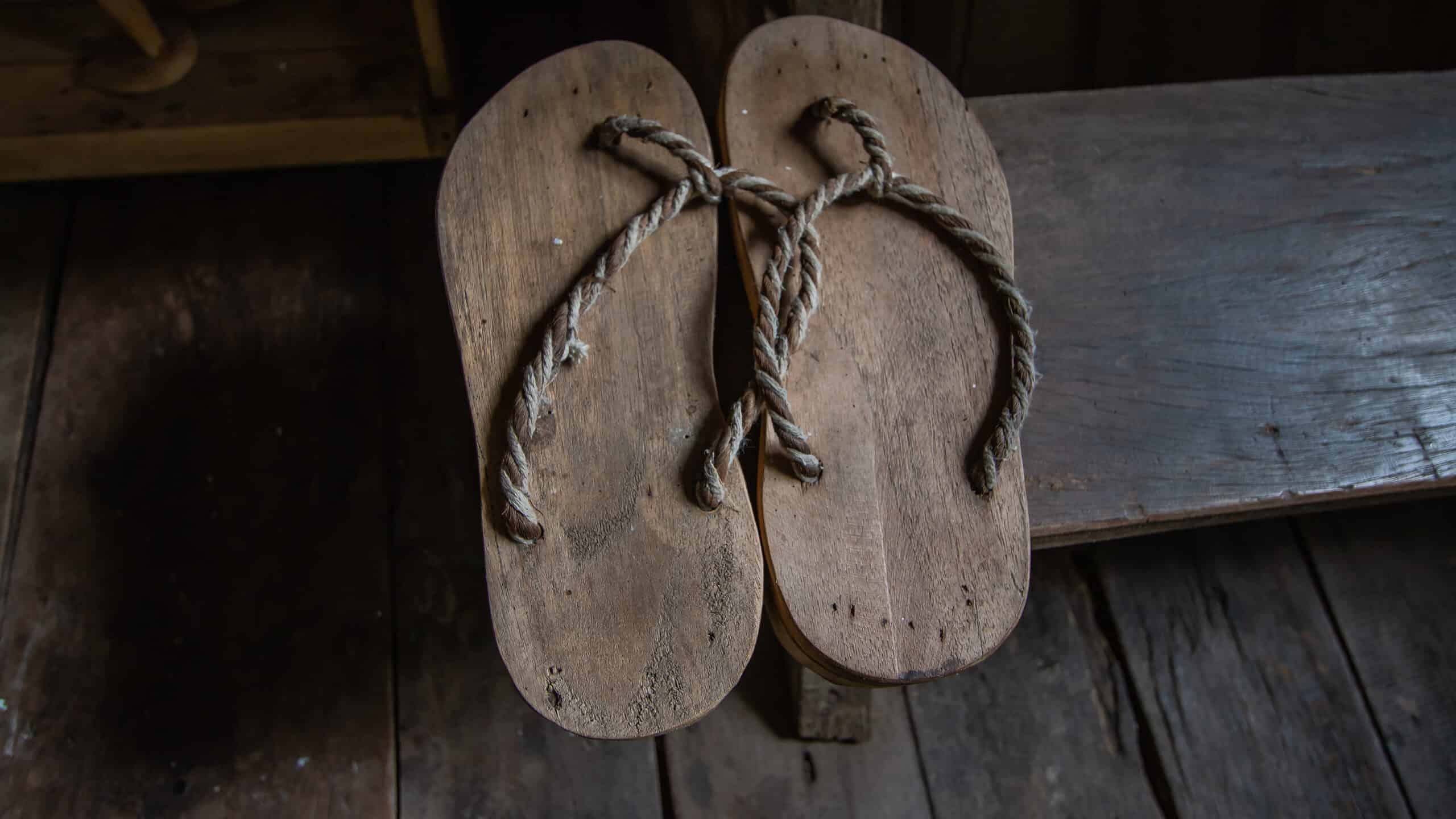 Center frame: Avery simple pair of rope and wooden sandals against a wooden background. The sandals resemble ancient flip-flops, with a loop of string attached to the wooden sole at the top for the toes to grasp, with one upside down V shape of rope attached at the heel end of the wooden sole on theft and the right. All natural colors.