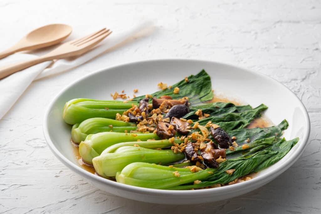 A white ceramic bowl of baby bok choy in an oyster sauce with Shitake Mushrooms and fried garlic. The bok choy has been cooked. Its leaves are darker green than its stems. the mushrooms and garlic are different shades of brown and gold. a brown sauce/juice is visible in the bottom of the bowl. It looks like soy sauce, but is oyster sauce. The bowl sits on a table covered with a white table cloth. a wooden (bamboo?) spoon and fork are resting on a napkin behind the bowl. The cloth covered table takes up the background.