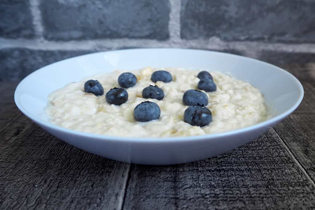 Alight gray ceramic rimless bowl of oatmeal, colored oatmeal, with 10 blueberry-blue blueberries arranged in a triangle, in 4 rows from left frame. the first row has 1 blueberry, the second row has 2 blueberries, the third row has 3 blue berried, and the fourth row has 4 blueberries. the bowl is center frame om an aged/weathered wood table with a blurry granite gray brick wall ib the background