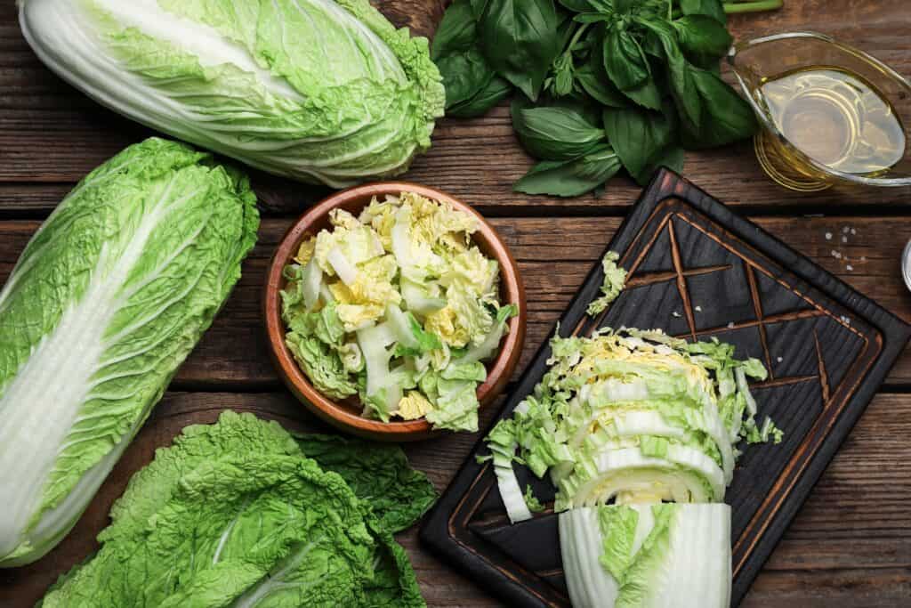 Overhead shot of (center frame) Chopped uplight green, dull yellow, and white Chinese cabbage in a wooden bowl, surrounded by, to the right, a 1/2 chopped head of Chinese cabbage.The cabbage is on a rectangular, wet wooden cutting board with diamond shapes carved into it. Above the cutting board is a clear glass vessel of golden, transparent olive oil, to the left of which are green basil leaves in a heap. Three full heads of Chinese cabbage take up the left part of the frame. All is setting on a natural wooden table made f planks