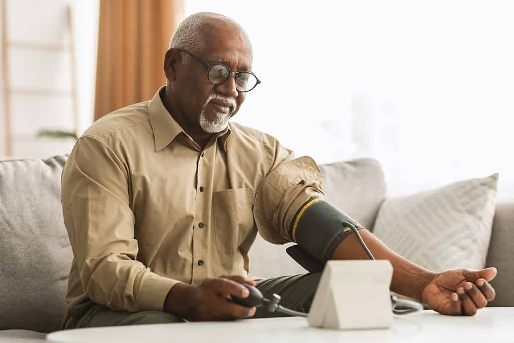 Frame left, a brown skinned man with  close-cropped grey hair and a gray van dyke, which is a mustache and chin whiskers . The man is seated a neutral colored textile covered  love seat - possibly a sofa. An end pillow, that matched the sofa is visible.. There is a white table in the foreground on which sits a blood pressure monitor. The man, who is wearing a button front collared khaki colored shirt, has a  black fabric /velcro blood pressure cuff wrapped around his left arm. His right hand is holding the pump that makes the cuff tight. The man is checking his blood pressure. A rust-colored drape panel is visible ibehind the man, otherwise white, indistinct background. 