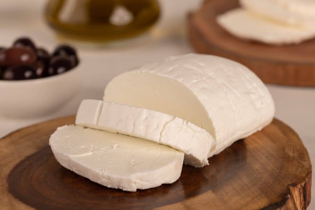 Center frame: Karish Cheese. a soft white cheese  on a natural slab of wood, with two generous slices cut from the oval shaped main cheese mass. The background consists of ripe black olives in a white ceramic bowl , frame left, and out of focus food at top of frame. 
