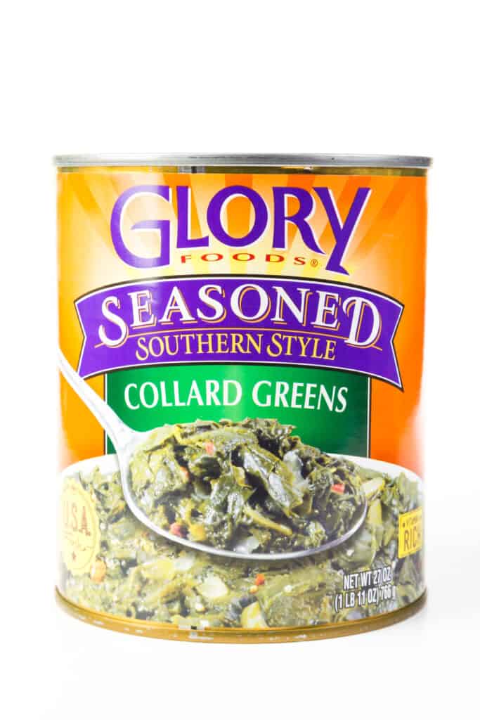 Can of Glory Foods Collard Greens seasoned Southern Style against white background. The can's label is colorful. The late is orange with green and purple highlights. At the bottom of the label is a big bowl of cooked green, with a large spoonful of greens  covering above the center of the bowl.