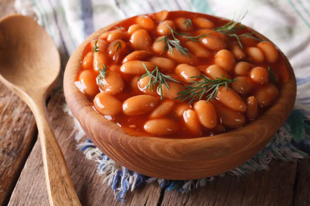 A small wooden, on a blue and white place mat, filled to the brim with orange/red baked beans with a couple of short sprigs of thyme. A wooden soon rest to the left of the bowl., off of the placemat. 